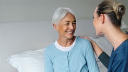 A smiling woman with gray hair sits on a bed next to a nurse in blue scrubs.