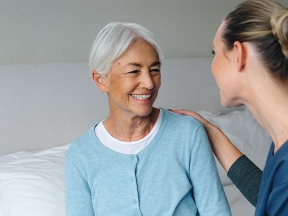 A smiling woman with gray hair sits on a bed next to a nurse in blue scrubs.