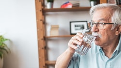 Elderly man with glasses drinks water near shelves and a potted plant by a window.
