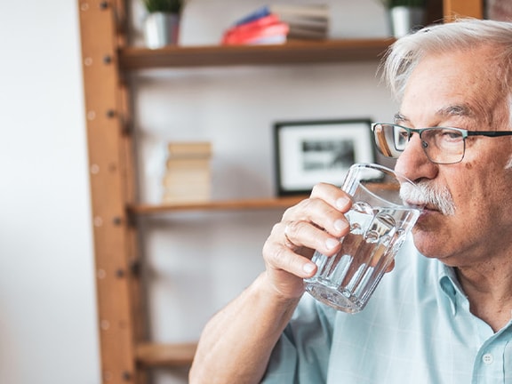Elderly man with glasses drinks water near shelves and a potted plant by a window.