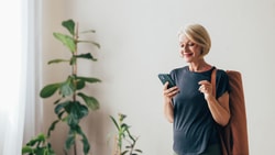 A woman with short hair holds a phone and yoga mat in a room with plants.