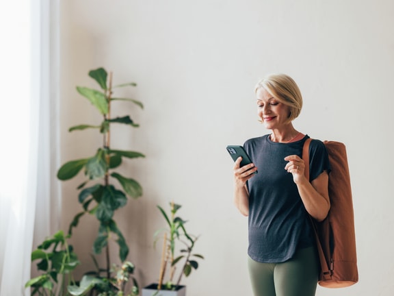 A woman with short hair holds a phone and yoga mat in a room with plants.