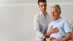 A young man helps an elderly man exercise in a well-lit room, promoting active living with medbridge.