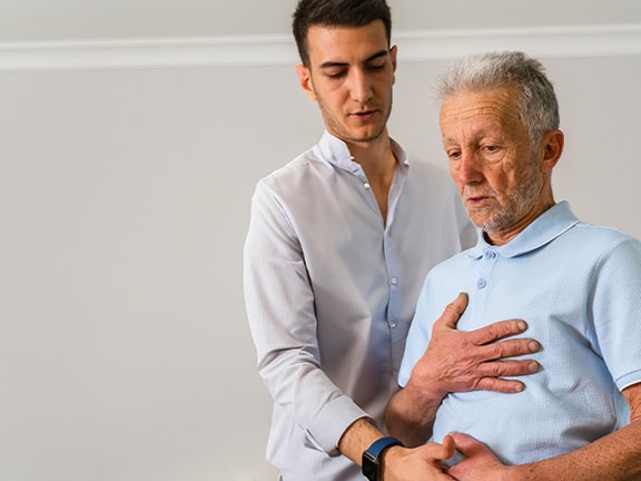 A young man helps an elderly man exercise in a well-lit room, promoting active living with medbridge.