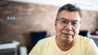An older man with glasses and gray hair in a yellow shirt sits indoors, reflecting on medbridge healthcare education.