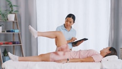 A physical therapist uses medbridge software while observing a woman lift one leg during a rehab session.