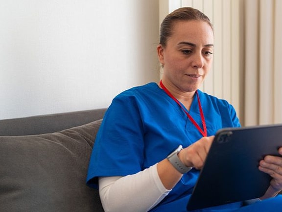 A healthcare professional in blue scrubs sits on a gray couch using a tablet for medbridge.