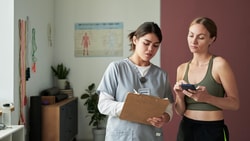 Clinician in scrubs holding a clipboard beside a patient in workout clothes checking a phone in a rehab clinic.