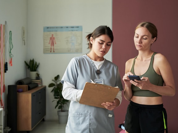 Clinician in scrubs holding a clipboard beside a patient in workout clothes checking a phone in a rehab clinic.