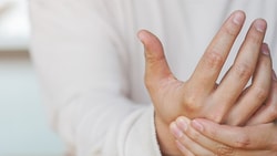 A person in a light shirt clasps their left hand, set against a plain backdrop.