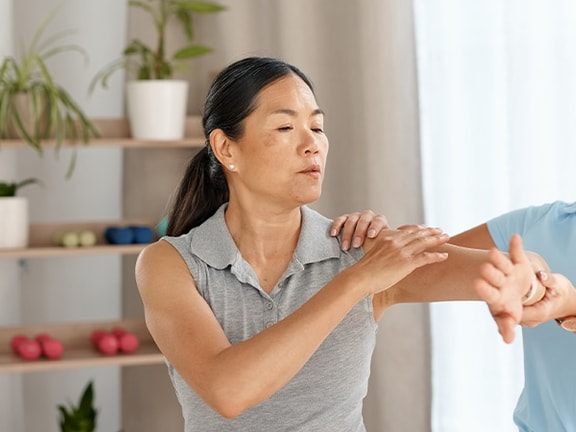 A woman helps another with an arm exercise in a Medbridge room with shelves and plants.