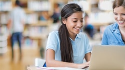 Two people at a library table smiling at a laptop, with bookshelves behind them.