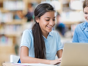 Two people at a library table smiling at a laptop, with bookshelves behind them.