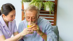 A medbridge caregiver helps an elderly man drink juice with potted plants in the background.