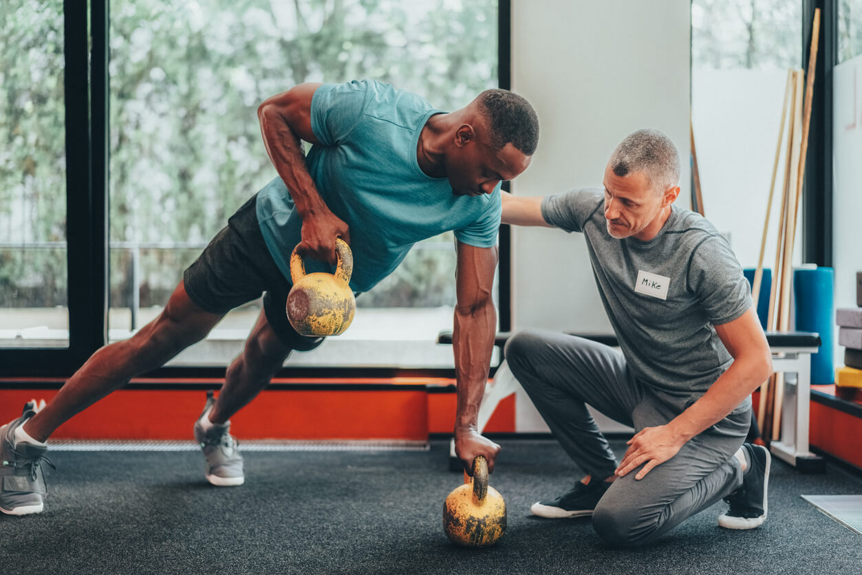 A man does a kettlebell row in a gym with a medbridge trainer kneeling beside him to provide support.