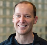 Smiling man with short brown hair in a black collared shirt, with a blurred background; professional at medbridge.