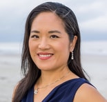 A woman with long dark hair and pearl earrings smiles on a beach, wearing a navy blue sleeveless top, embodying medbridge warmth.