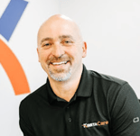 Smiling bald man with a goatee in a black "Kinetacare" polo, sitting indoors with a white wall and colorful background.