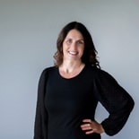 Smiling woman with wavy dark hair in black top stands in front of a light gray background, representing medbridge.