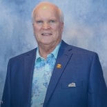 An older man in a blue suit and patterned shirt stands before a blue-gray backdrop, representing medbridge professionalism.