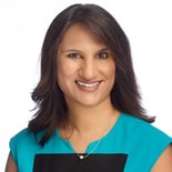 Smiling woman with shoulder-length dark hair in blue and black top and necklace, standing before a plain white background.