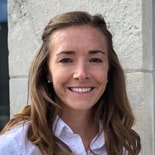 A woman with long brown hair in a white shirt smiles in front of a stone wall, representing medbridge’s positive impact.