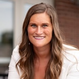 A woman with long wavy brown hair in a white blazer smiles confidently outdoors, representing medbridge healthcare training.