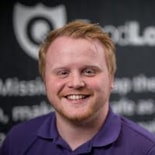 A smiling person with short red hair and a beard stands before a blurred medbridge shield logo and white text.