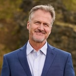 A confident, middle-aged man in a blue suit stands outside by rocks, representing medbridge digital healthcare professionals.