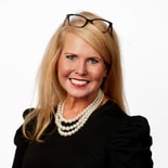 A blonde woman in black glasses and pearls smiles at the camera, representing medbridge's digital healthcare.