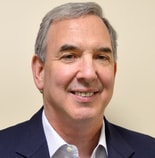 A smiling middle-aged man with gray hair and a blazer stands in front of a plain background, representing medbridge.