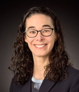Person with long curly brown hair, glasses, and a dark blazer smiles in front of a dark background, representing medbridge.