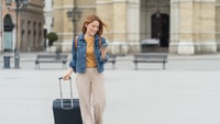 Travel therapist with suitcase using a phone to manage licensure and CEU requirements in a city plaza.