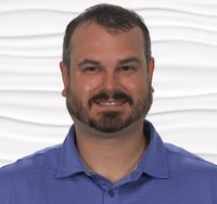 A bearded man in a blue polo shirt smiles against a wavy, white background.