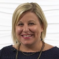 A smiling person with short blonde hair, in a navy blue top and beaded necklace, embodies professionalism in Medbridge healthcare training.