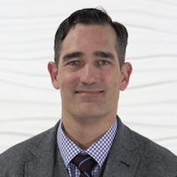 Man in a gray suit with a checkered shirt and striped tie, set against a textured white backdrop for medbridge.