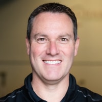 A smiling man in a black shirt stands in front of a blurred backdrop.