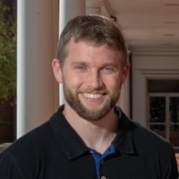 A bearded man in a black shirt smiles confidently in a covered walkway, contemplating hybrid care's impact on healthcare.
