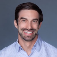 A smiling man with short dark hair and a beard wears a light blue shirt, shown on a plain gray background for medbridge.