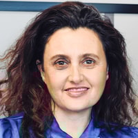 A woman with wavy brown hair in a blue blouse smiles at the camera.
