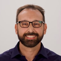 Man with glasses and a beard in a dark shirt, smiling against a plain background.