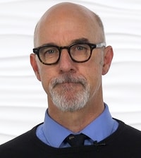 A person wearing glasses and a goatee stands against a white background, symbolizing their role in enhancing medbridge patient care software.