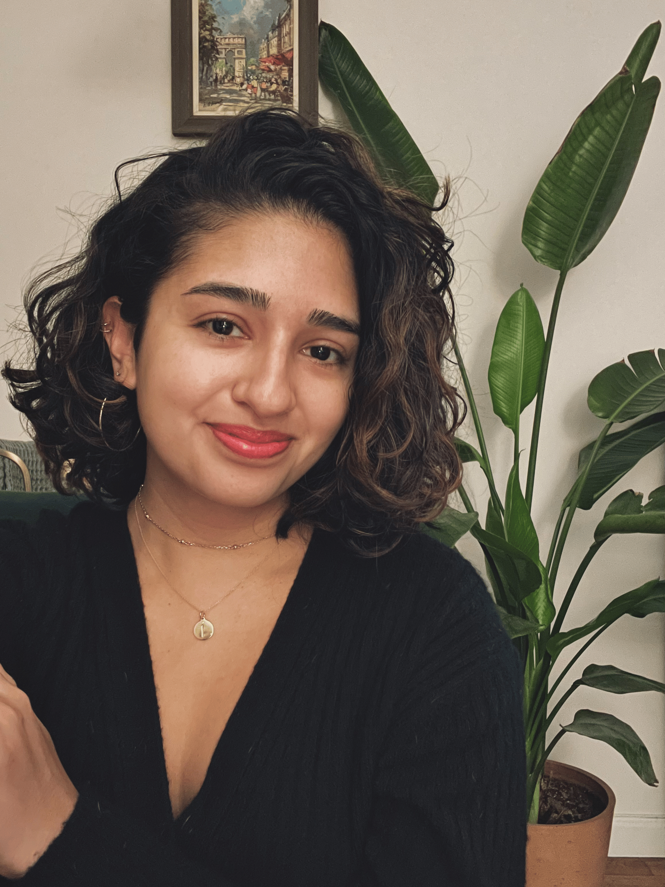 Smiling woman with curly hair and gold jewelry, in black top, sits near a green plant and art, reflecting her digital healthcare focus.