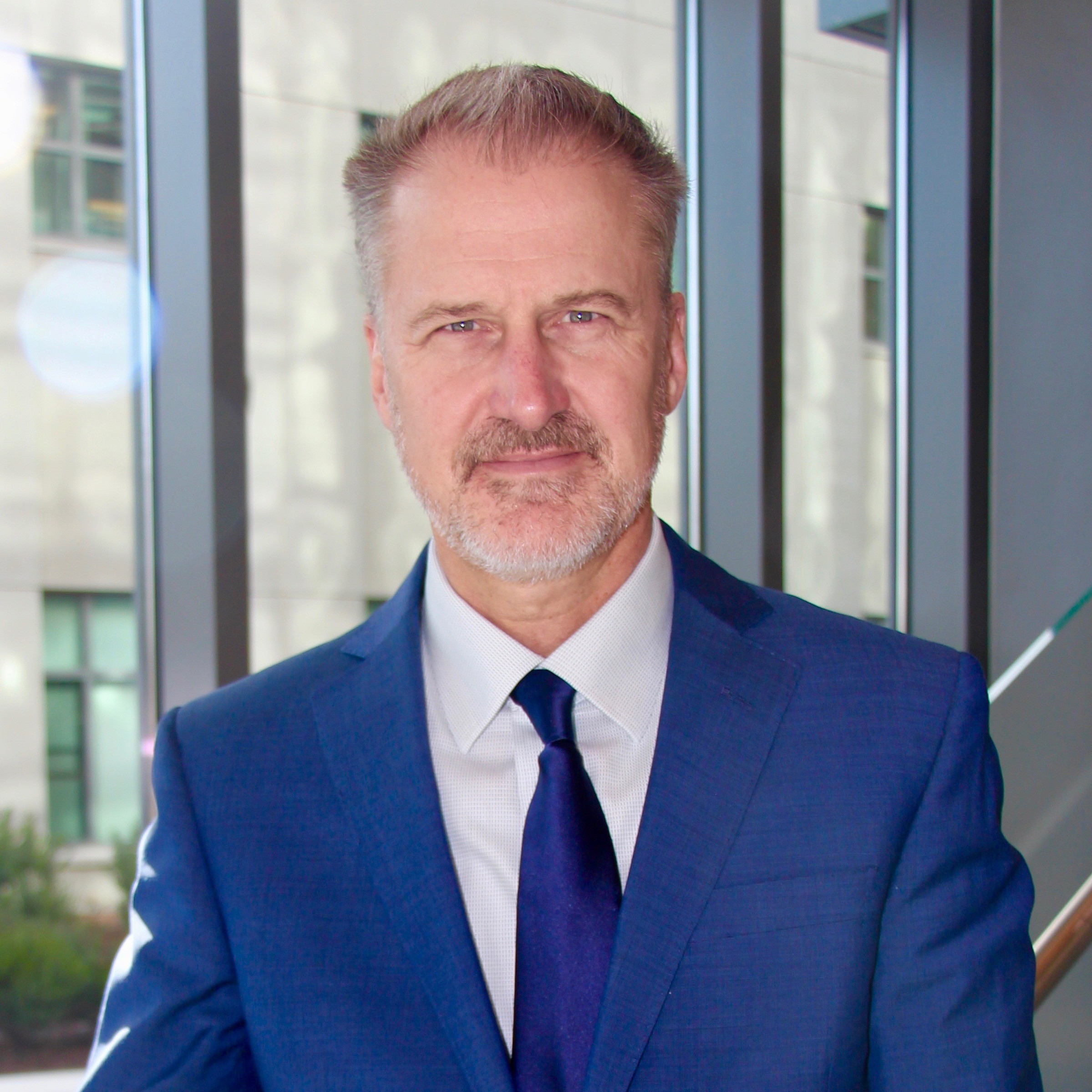 A gray-haired man in a blue suit and tie stands by large windows at medbridge.