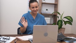 Clinician conducting remote therapeutic monitoring (RTM) telehealth visit, waving during video call on laptop in clinic office