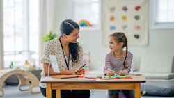 A smiling SLP and young girl engage in a play-based autism assessment at a small wooden table.