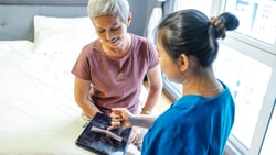 A healthcare worker shows patient info on a tablet to an older woman sitting on a bed; screen displays medbridge software.