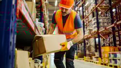 Worker in orange safety vest and helmet lifts a box in warehouse supporting medbridge healthcare training and hybrid care.