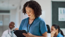 Nurse reviewing a tablet while completing a nursing skills checklist in a clinical setting.