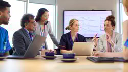 Healthcare professionals in a meeting discussing PEPM vs PMPM strategies using data charts on a screen.